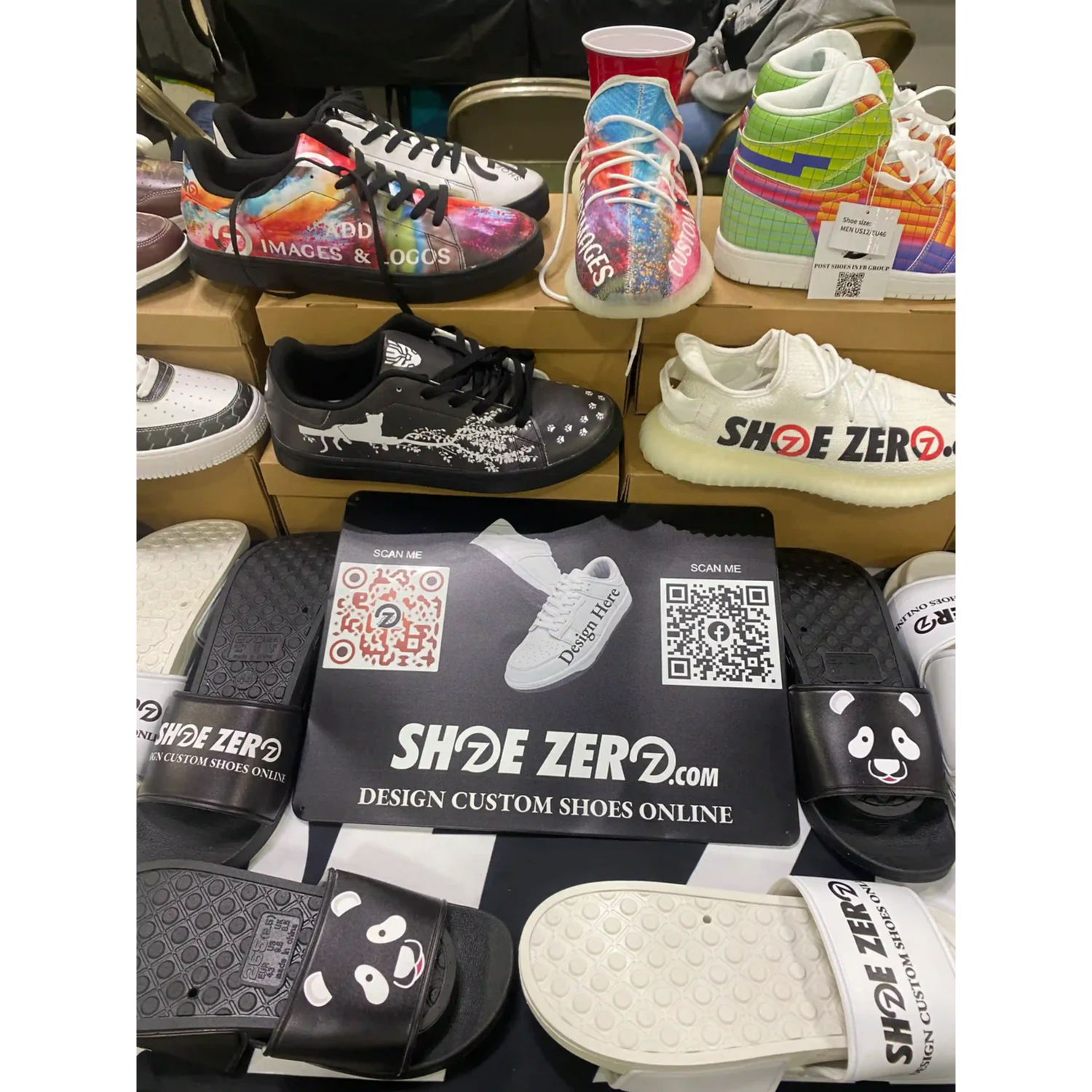 A display of custom-designed shoes and sandals atop cardboard boxes. Brightly colored, and black-and-white styles. A “Shoe Zero” poster with QR codes visible.