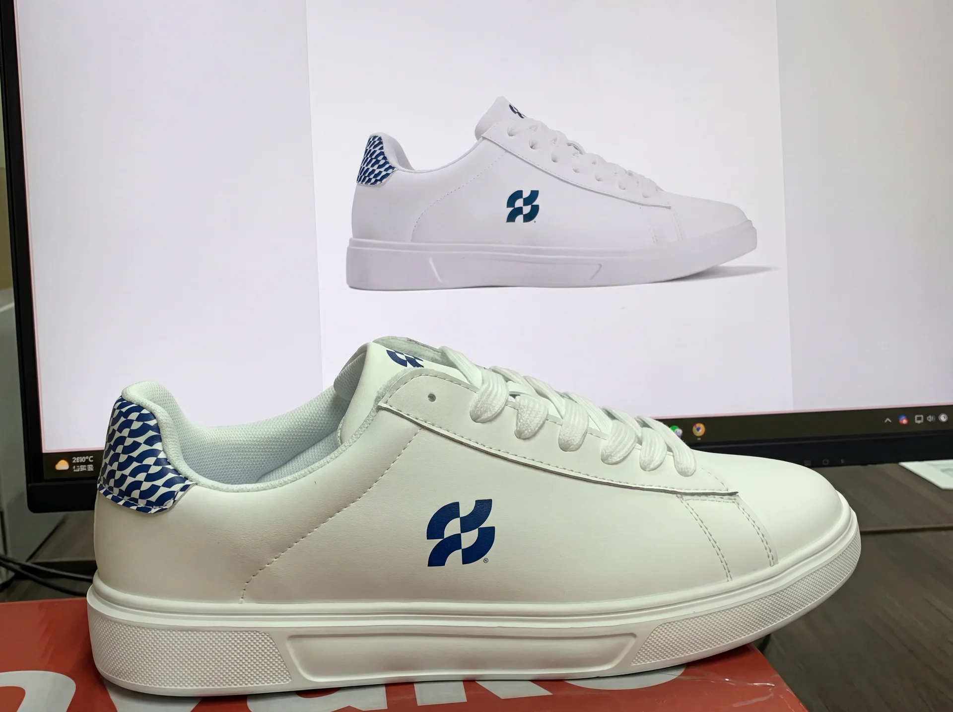 A finished custom Shoe Zero sneaker in white synthetic leather with a blue brand logo and blue patterned heel tab, sitting on its branded box with the design reference on the monitor behind